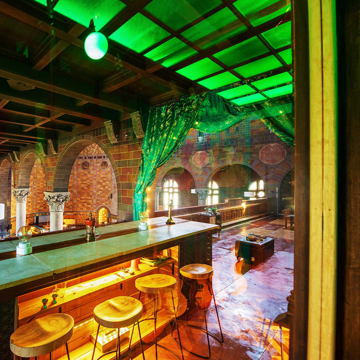 A brightly lit bar area with colorful tile work and green decorative elements, featuring wooden stools and various lamps.