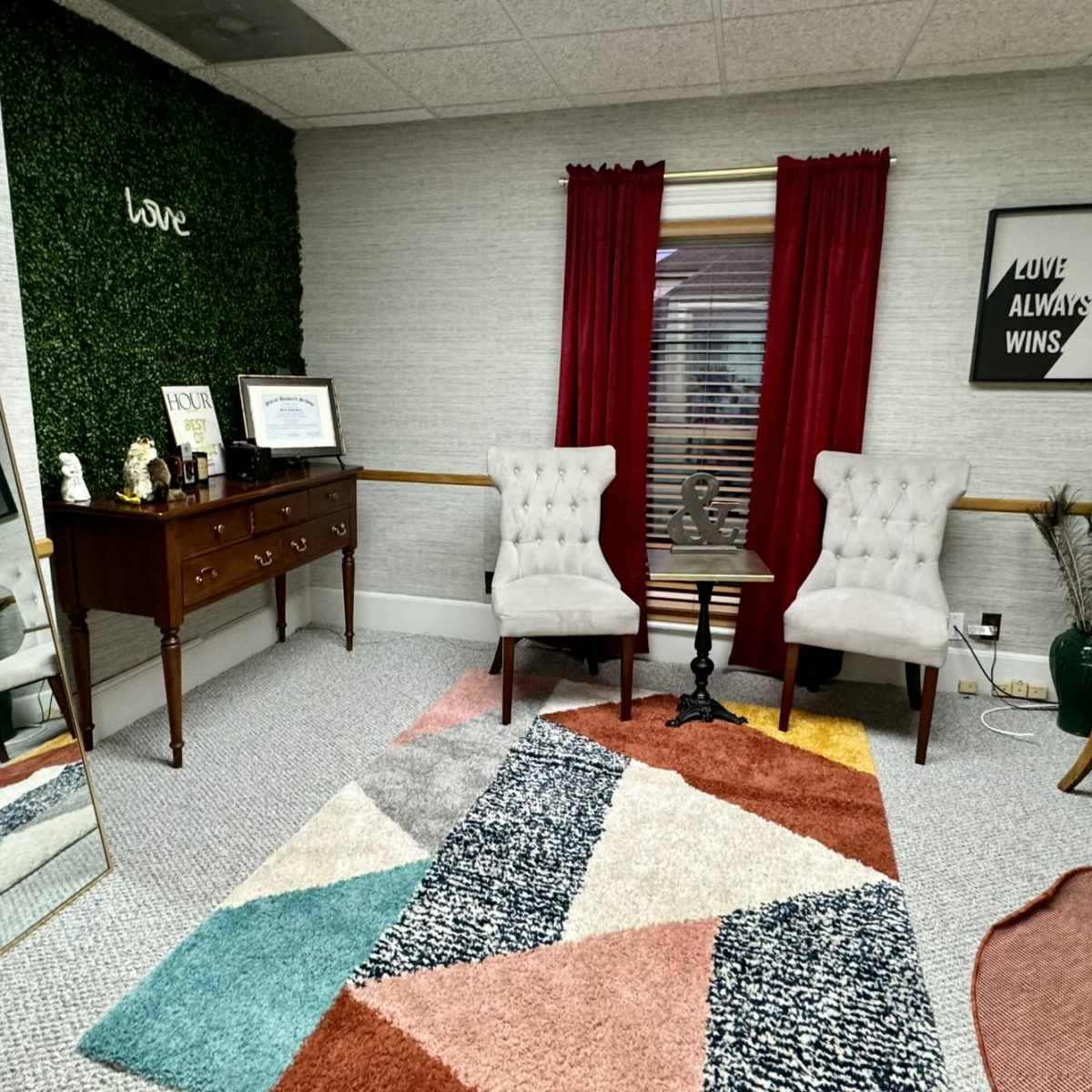 The room features a large mirror, two upholstered chairs, a wooden dresser, and a colorful geometric rug.