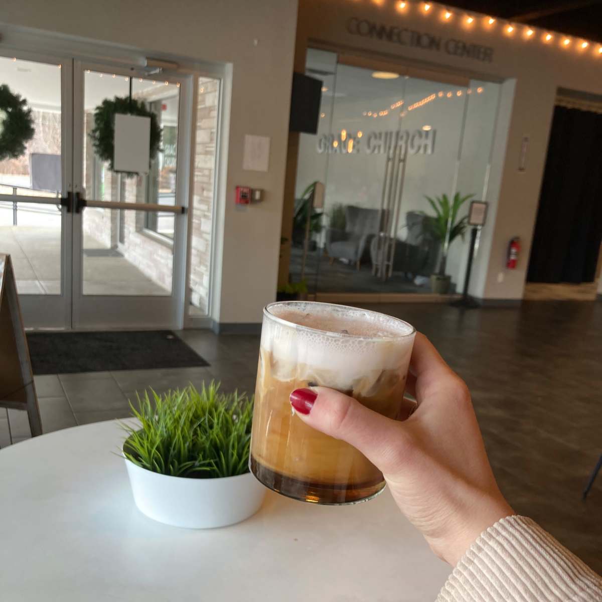 A person holds a glass of iced coffee in a modern interior space with a potted plant on a white table.