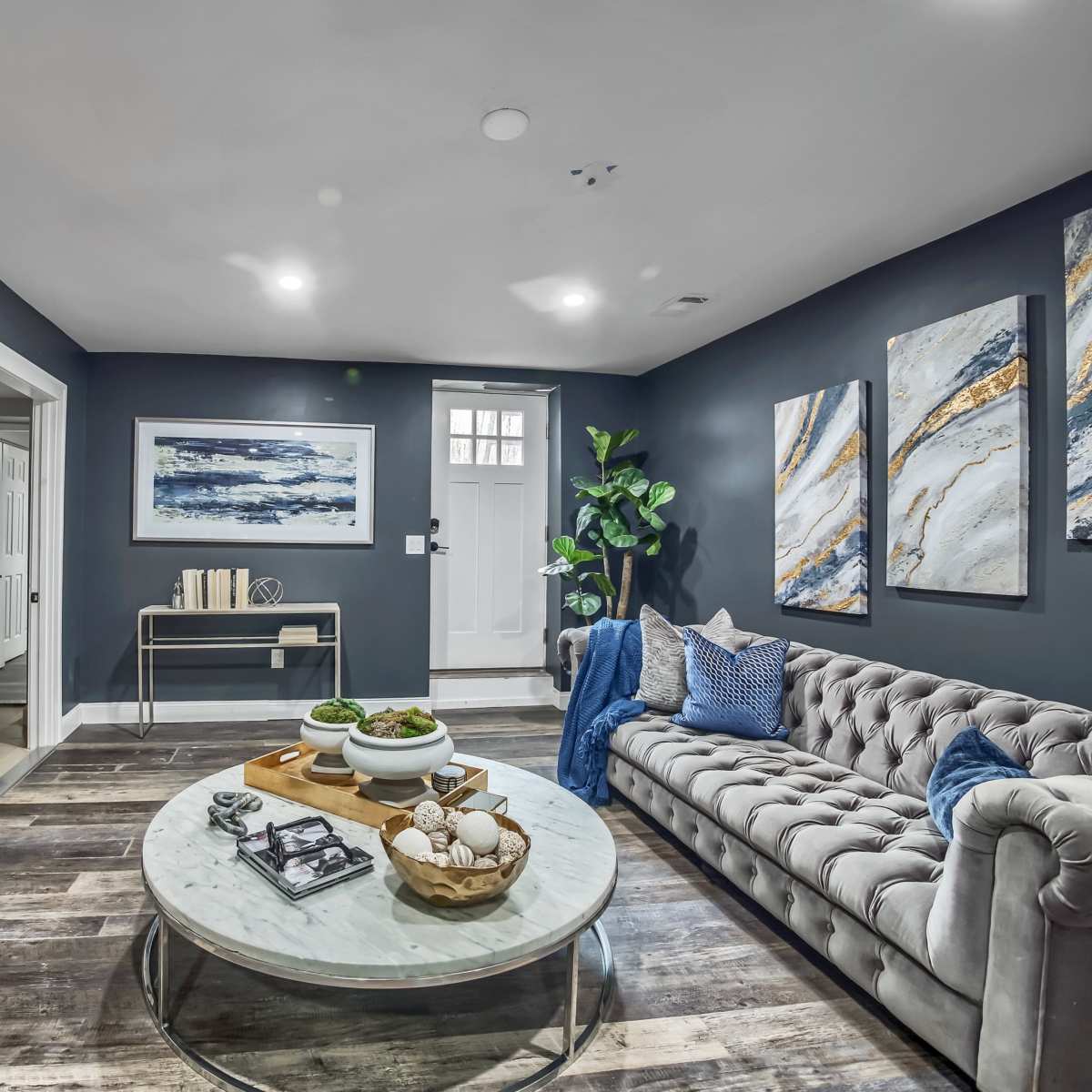 A modern living room with a gray tufted sofa, a round marble coffee table, a wall-mounted television, and an electric fireplace beneath a set of abstract paintings.