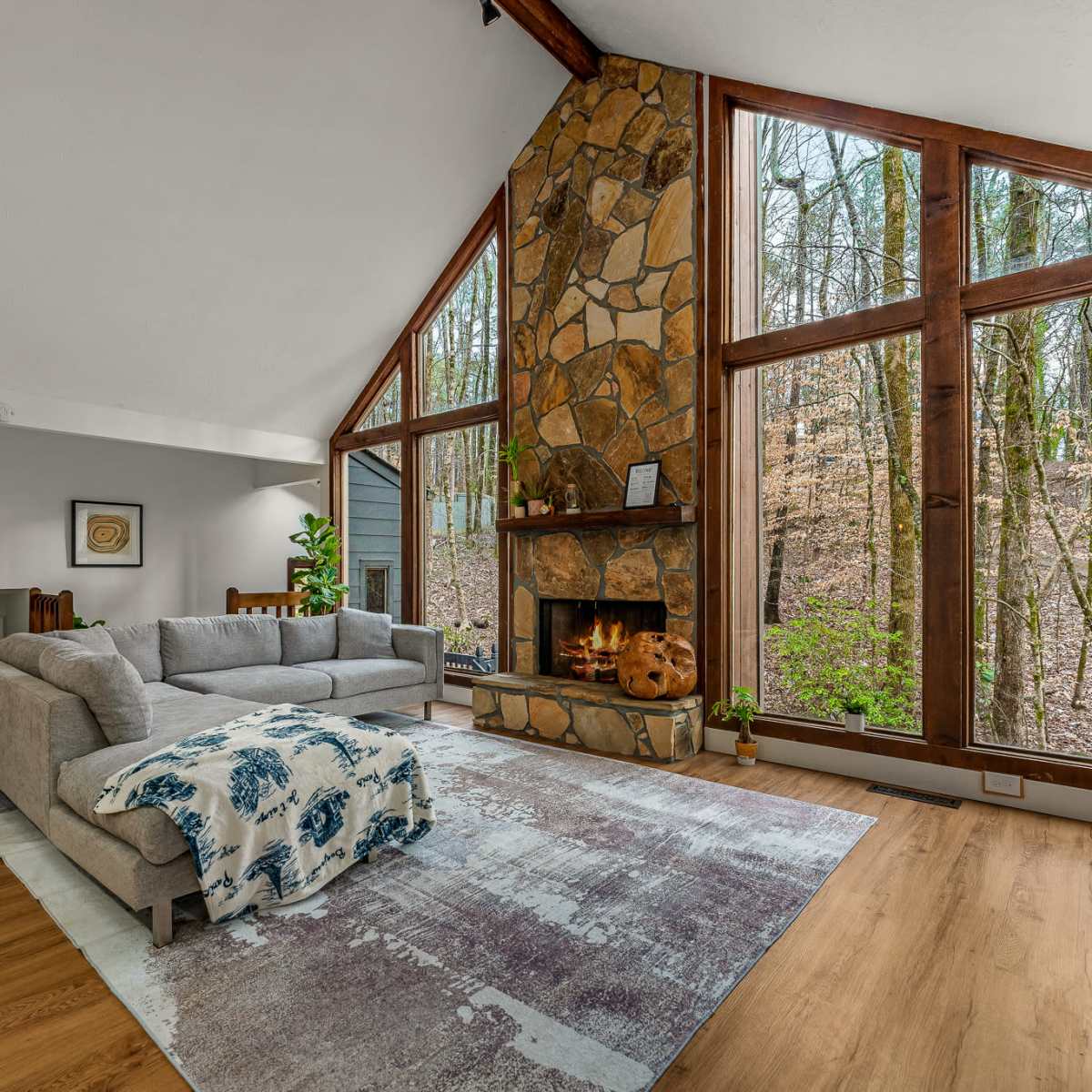 A modern living room features a large gray couch, a stone fireplace, and tall windows overlooking a wooded area.