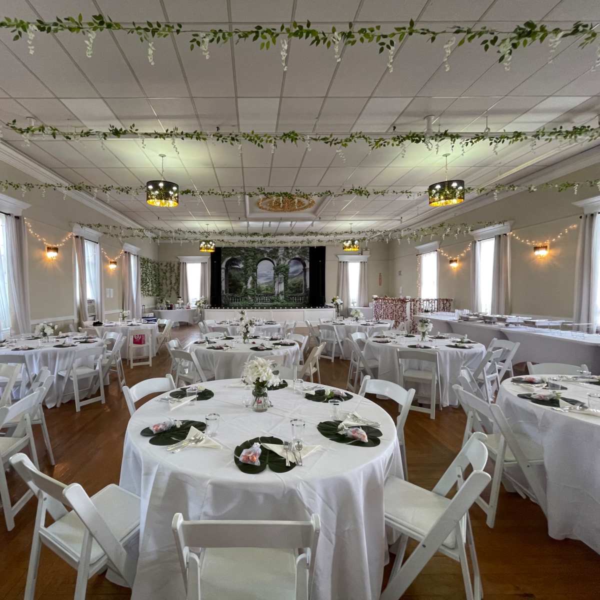 A banquet hall set up for an event, with round tables adorned with centerpieces and white chairs, all arranged under decorative greenery and lighting.