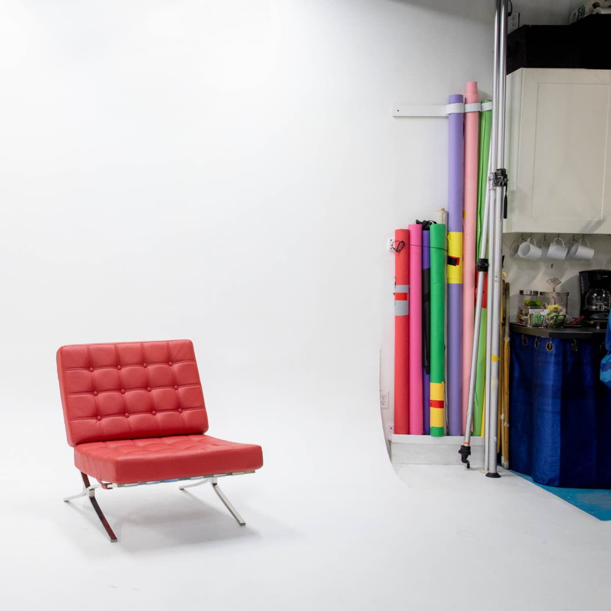 A red chair with a tufted design is positioned in the center of a photography studio with colorful backdrop rolls stacked to one side.