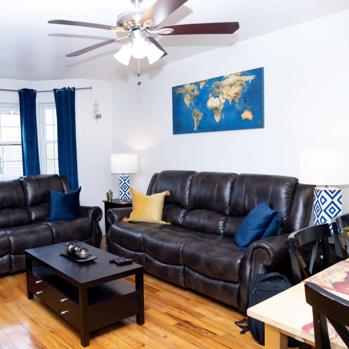 A living room features two brown leather sofas, a black coffee table, and a world map on the wall, with large windows dressed in blue curtains.