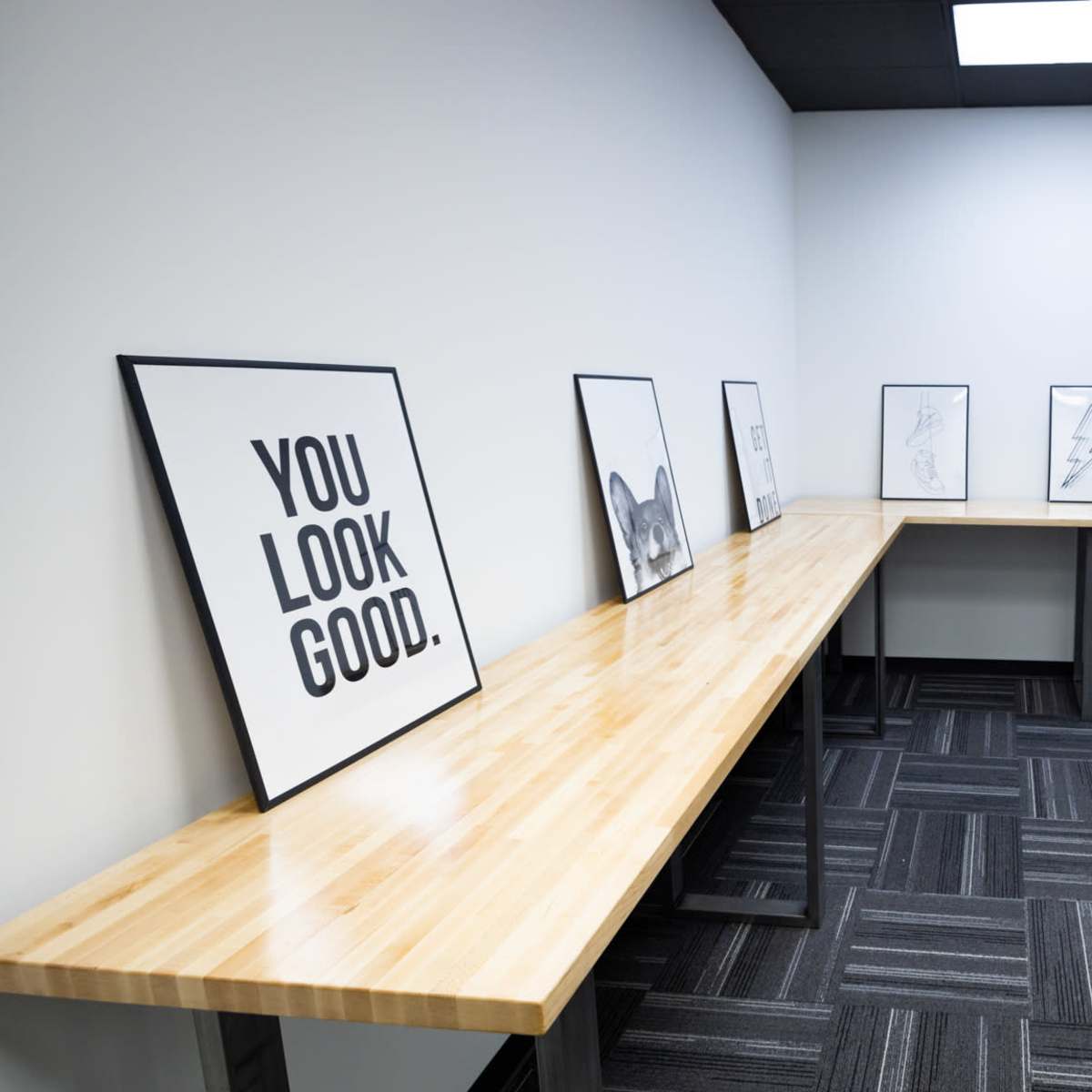 A minimalistic room features a long wooden table against a wall, displaying various framed posters with bold black text and designs.