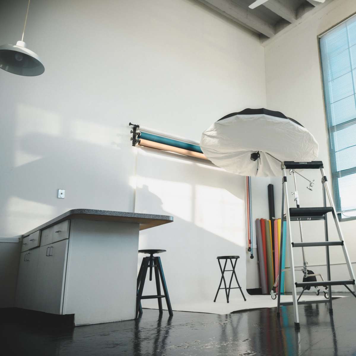 A photography studio with a large umbrella softbox, a ladder, a stool, and colorful backdrop rolls near a large window.