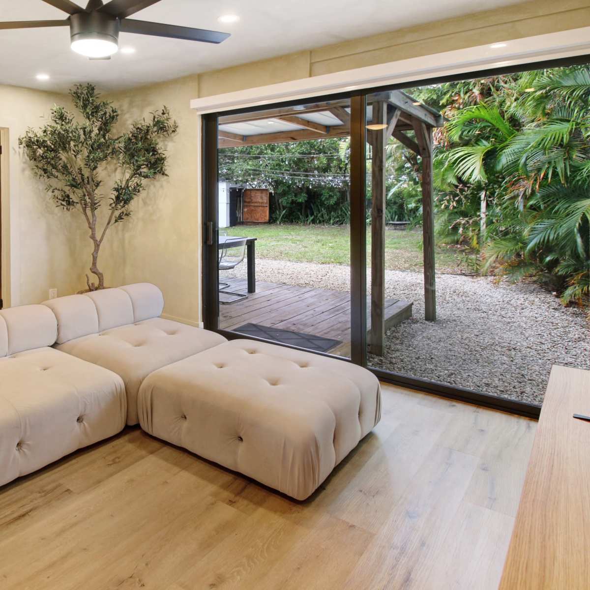 A beige tufted sectional sofa faces a large glass door that opens to a backyard with tropical vegetation and a wooden deck.