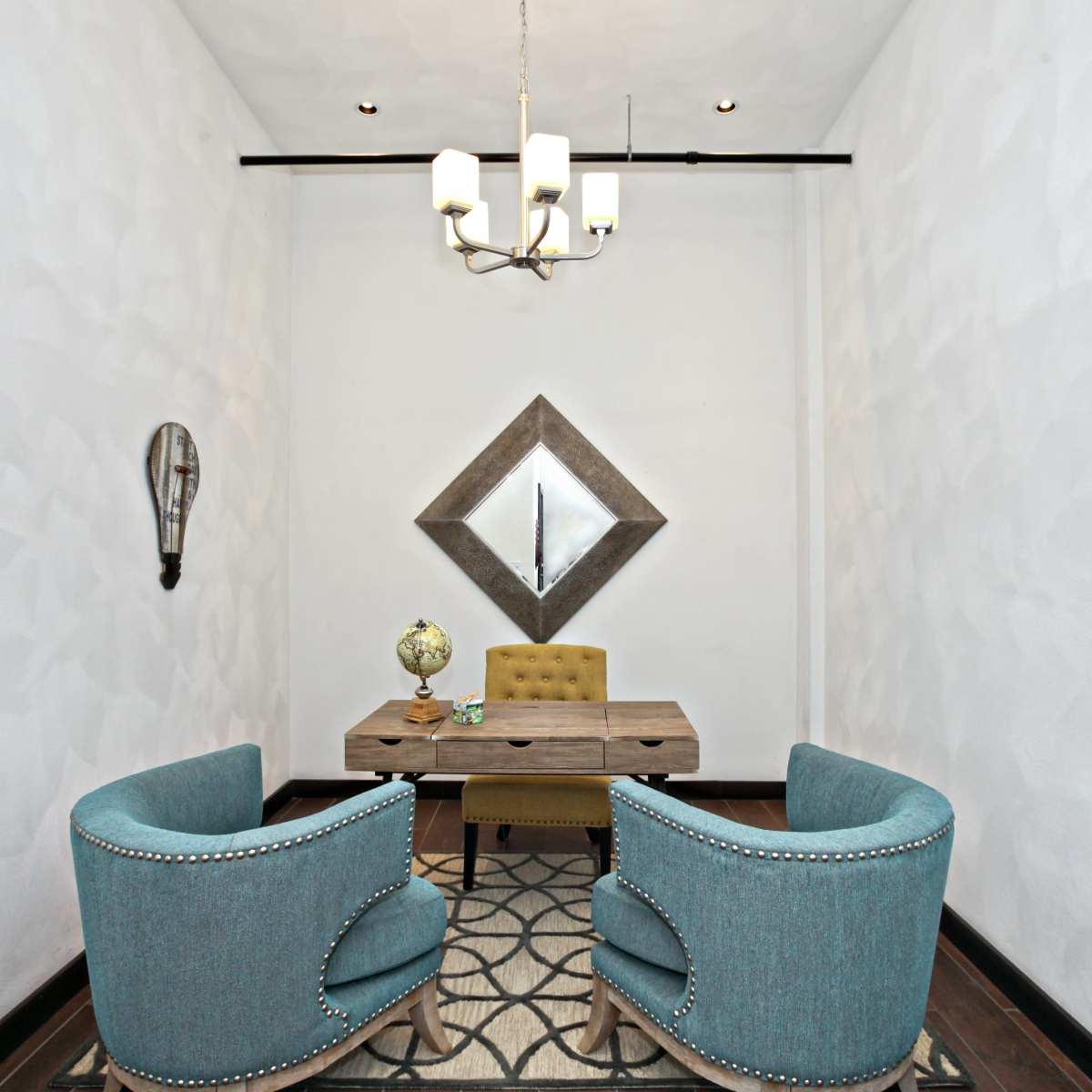A small room features two blue chairs and a wooden table under a chandelier, with a mirrored diamond hanging on the wall.