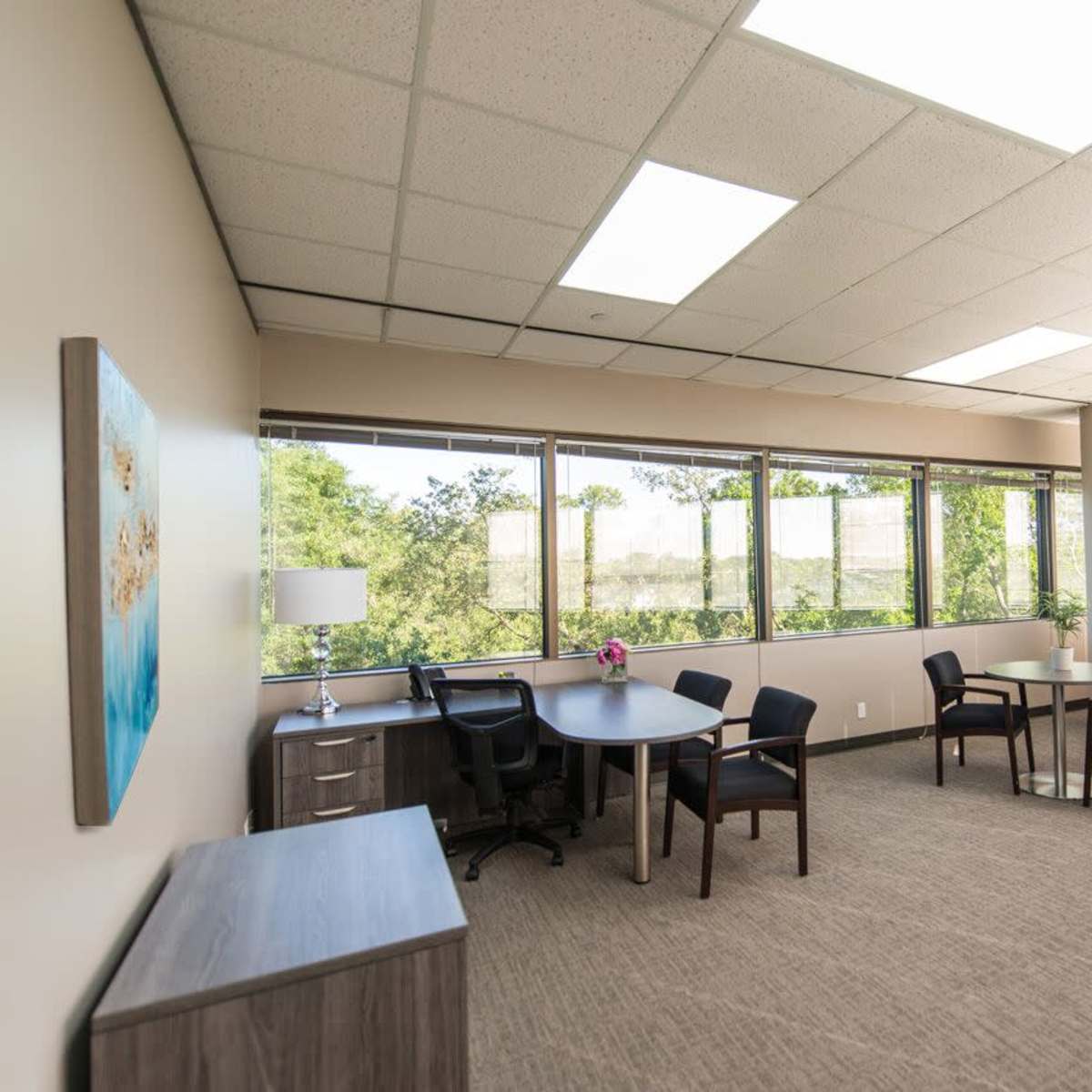 A modern office space with a round table, four chairs, and large windows allowing natural light to enter.