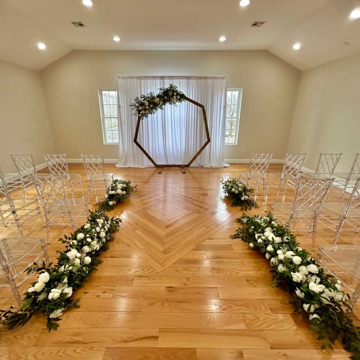 A wedding ceremony setup features transparent chairs arranged in a circular pattern around a wooden arch adorned with greenery, with white flowers lining the aisle.