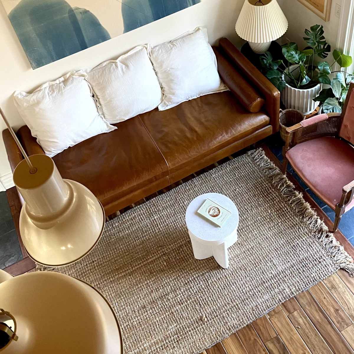 A cozy living room with a brown leather sofa, white cushions, a small white side table, and various plants, all under a wooden ceiling.