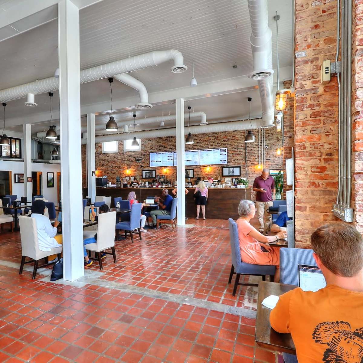 A spacious café with a mix of wooden and brick walls, featuring various seating arrangements and a counter in the background where staff interact with customers.