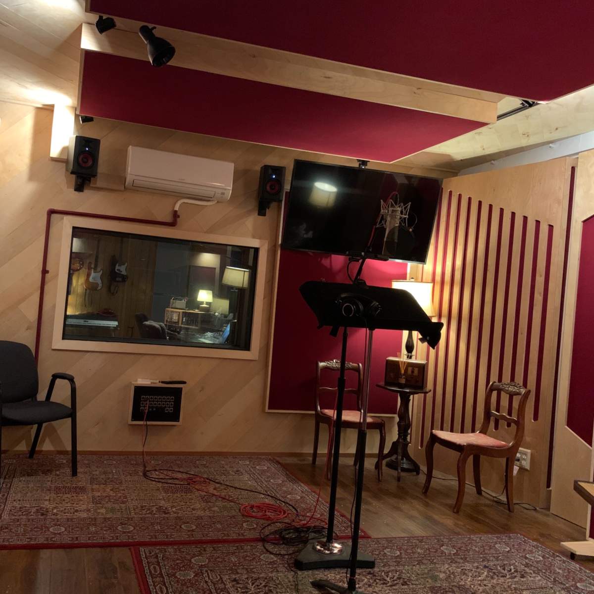 A recording studio with soundproofed walls, a control room visible through a window, and various seating arrangements including a chair and a stand for scripts.
