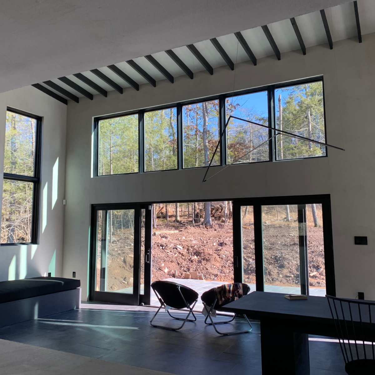 The interior of a modern room features large windows overlooking a wooded landscape, with a black couch and two lounge chairs positioned nearby.