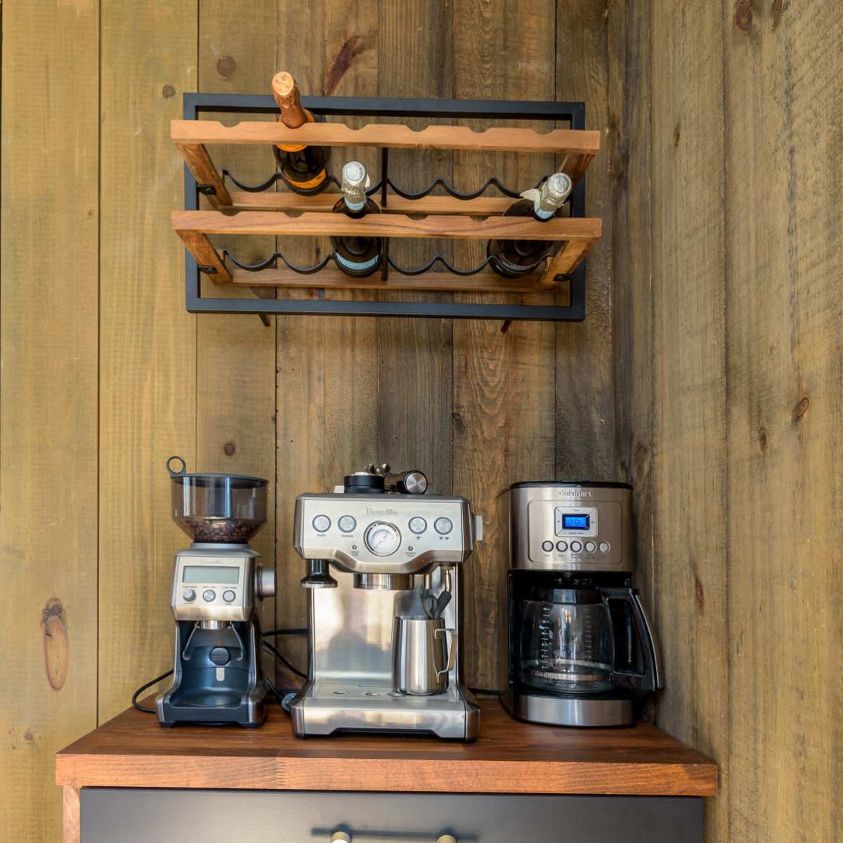 A coffee station with a grinder, espresso machine, and drip coffee maker arranged on a dark cabinet beneath a shelf displaying wine bottles.