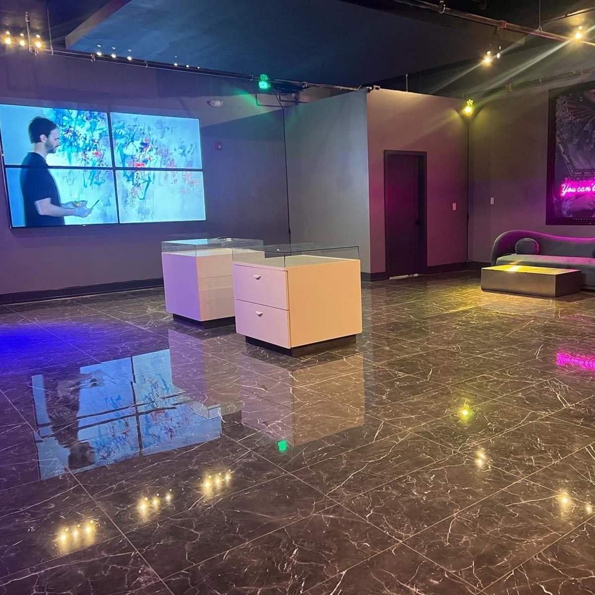 A modern gallery space with polished dark marble flooring, featuring a large screen displaying artwork, two white display cases, and a purple couch against the wall.