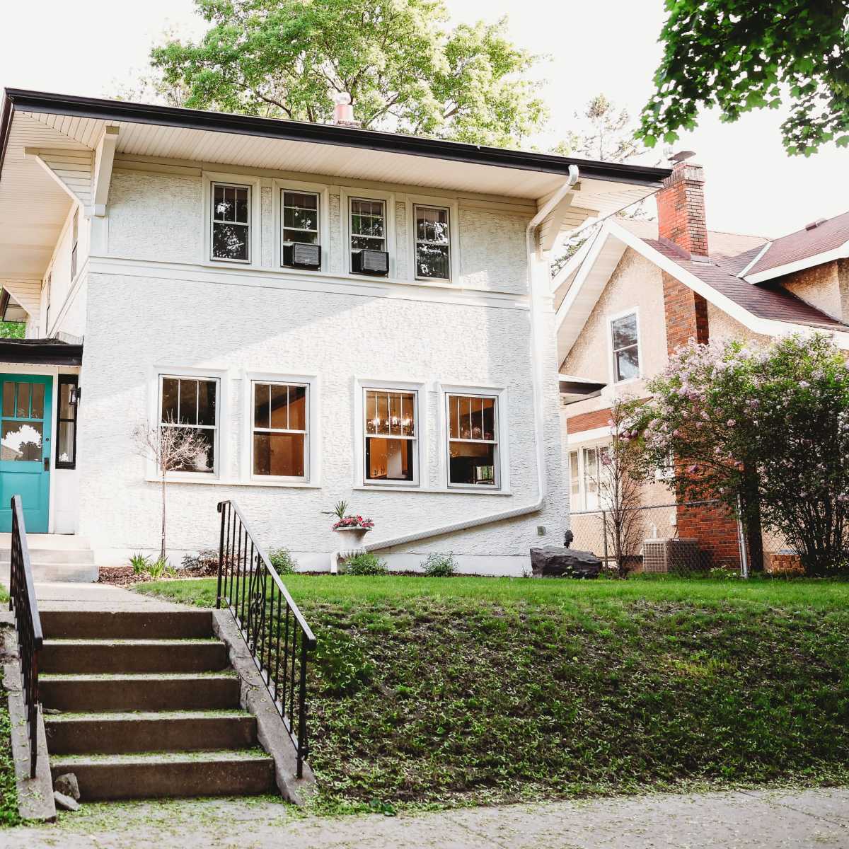 A two-story white house with a turquoise door and several windows is situated on a grassy lawn beside a tree-lined street.