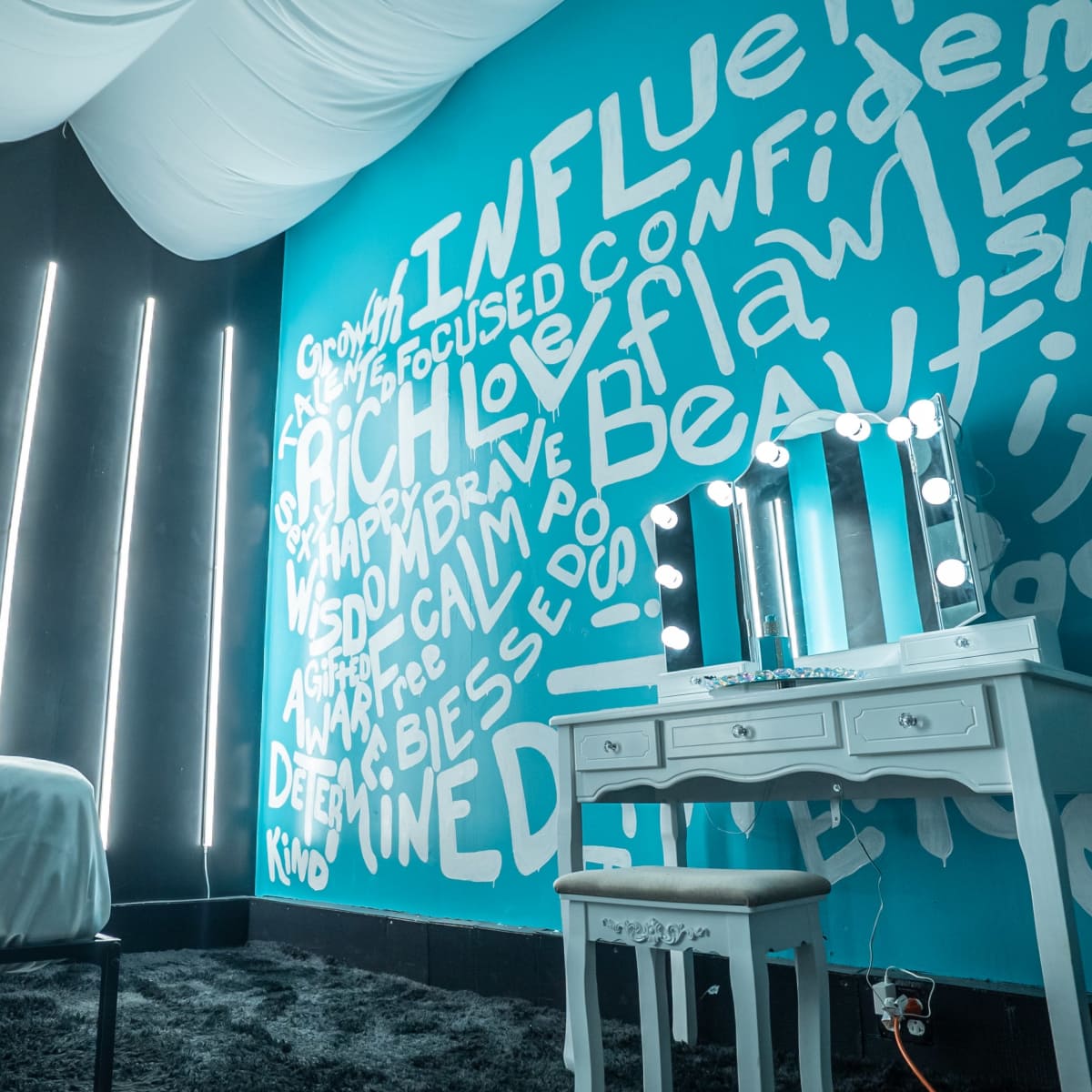 A modern bedroom with a white bed, a vanity table with mirrors and lights, and a feature wall covered in various inspirational words and phrases.