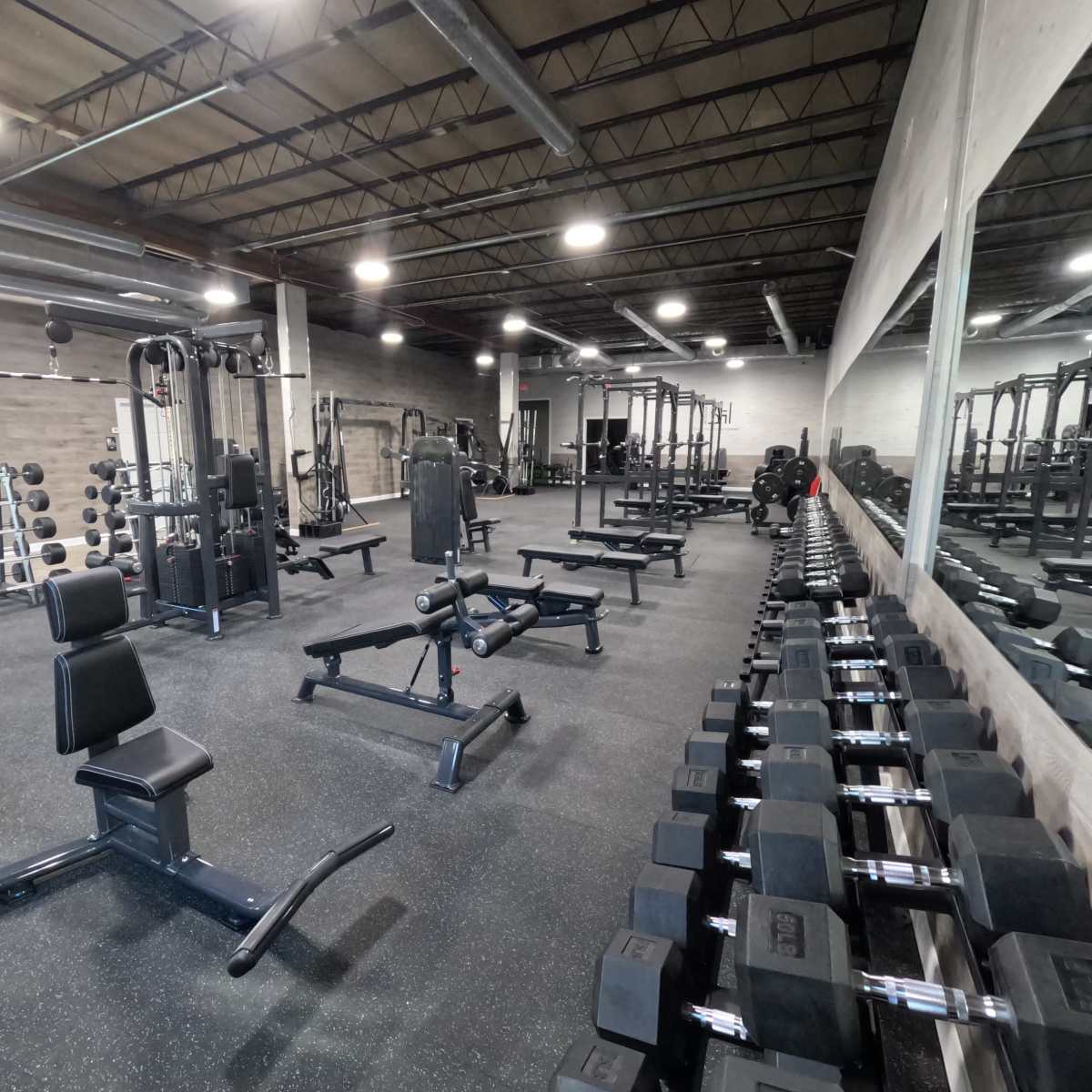 A spacious gym interior with various weightlifting equipment, including a row of dumbbells and benches, along with weight machines arranged across the room.