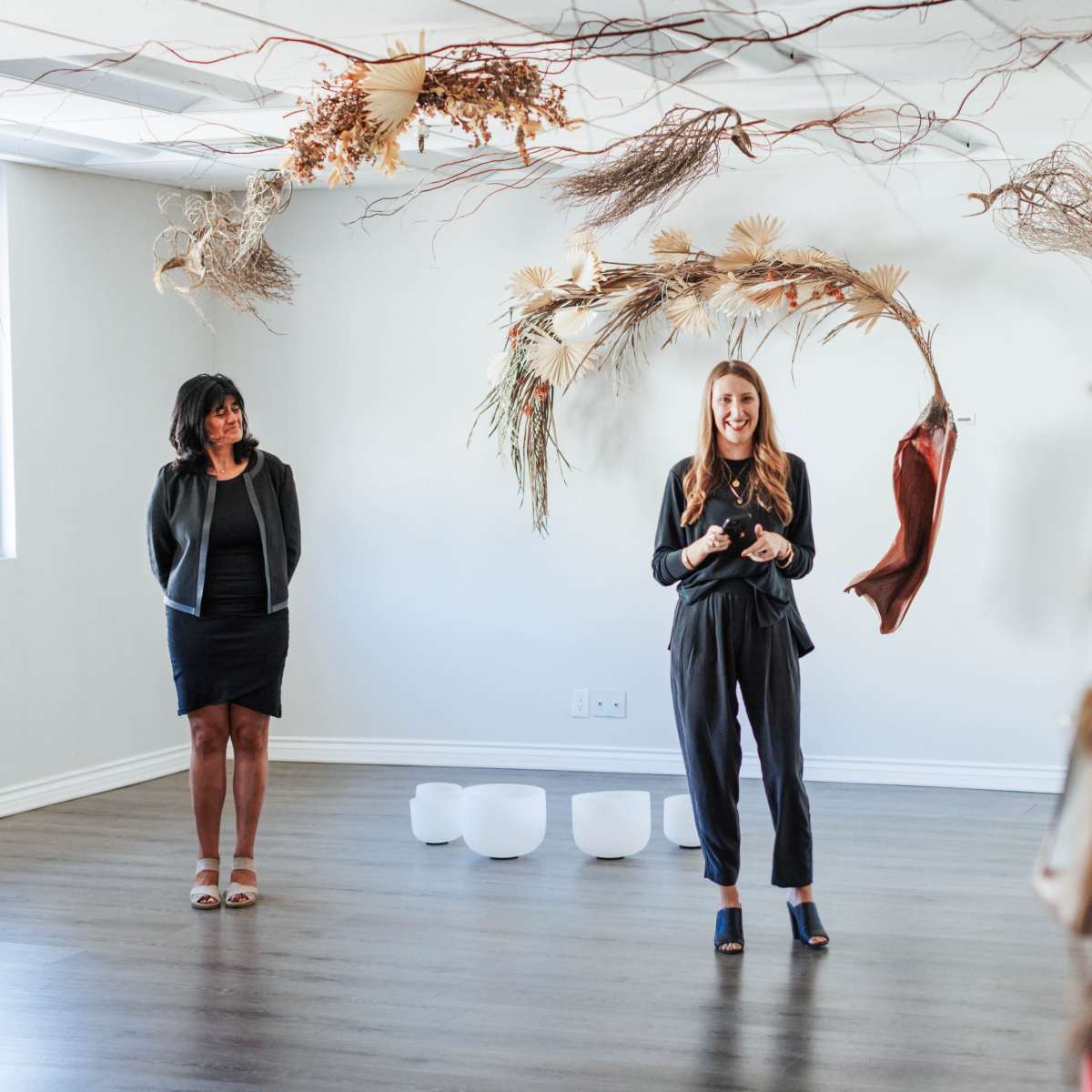 A woman in a black outfit stands speaking in front of a small audience, while another woman stands to her left, with decorative branches and materials hanging from the ceiling.