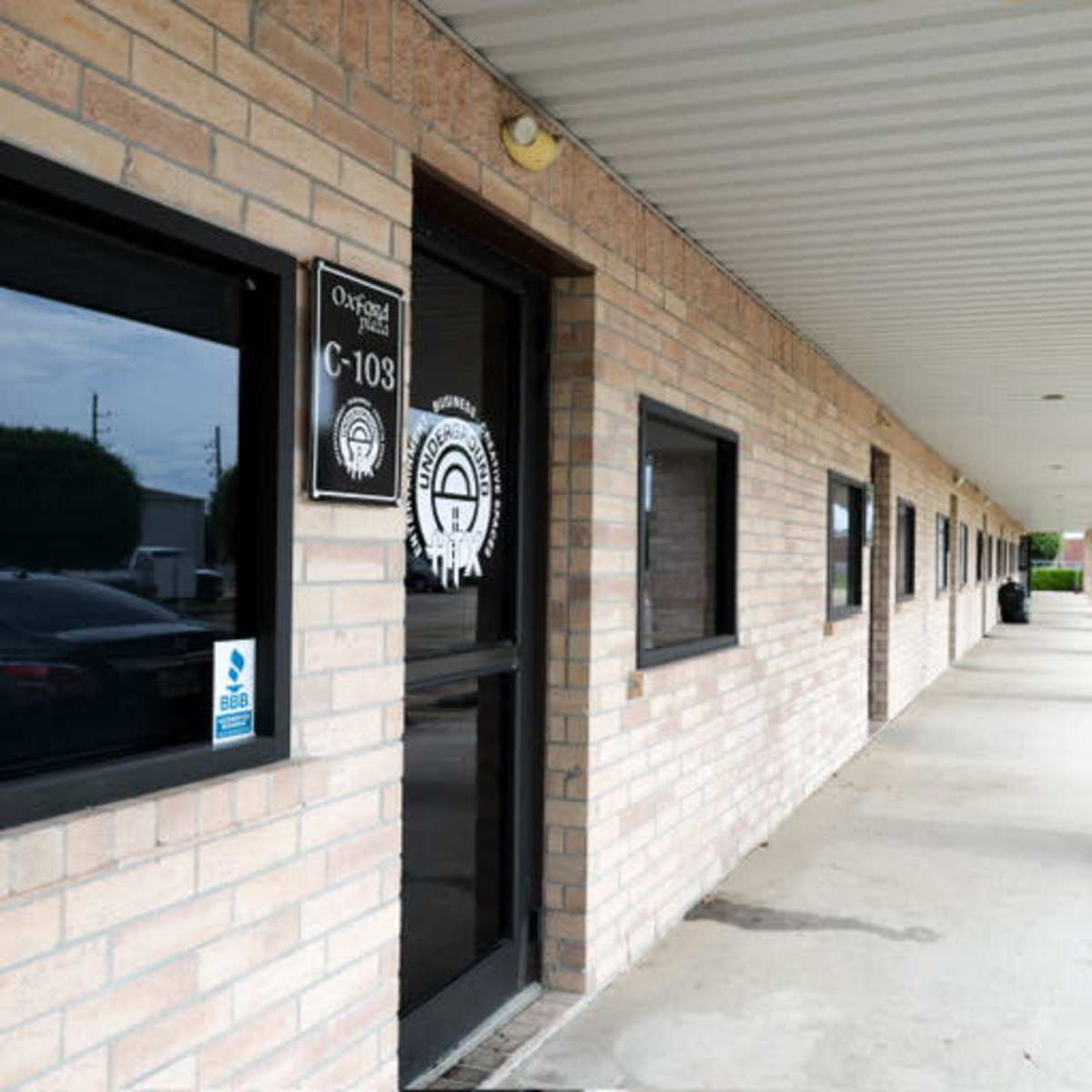 A brick building with a covered walkway and multiple windows, featuring a door labeled "C-103."