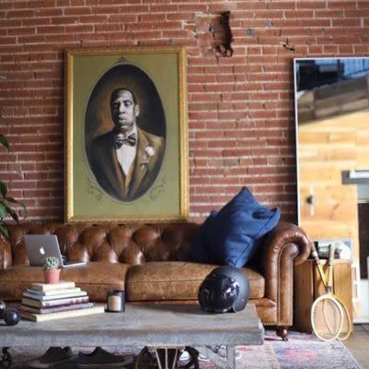A leather sofa against a brick wall, with a portrait of a man in formal attire hanging above it, and a rustic coffee table adorned with books and a decorative item.