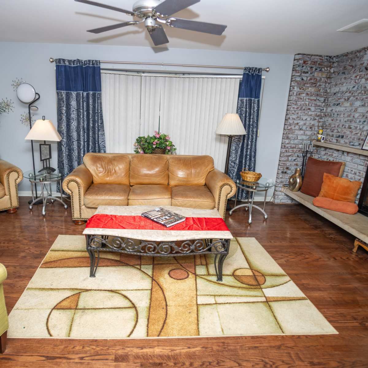 The living room features a mix of seating, including tan leather sofas and a green chair, arranged around a coffee table on a patterned area rug, with a fireplace and decorative elements in the background.