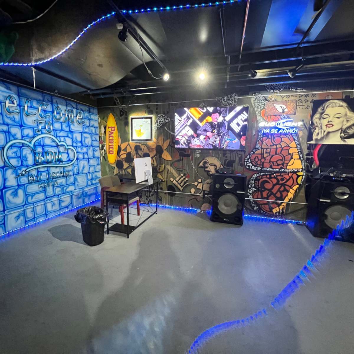 An underground room with colorful graffiti on the walls, a table and a trash can on the floor, and large speakers positioned near a television screen.