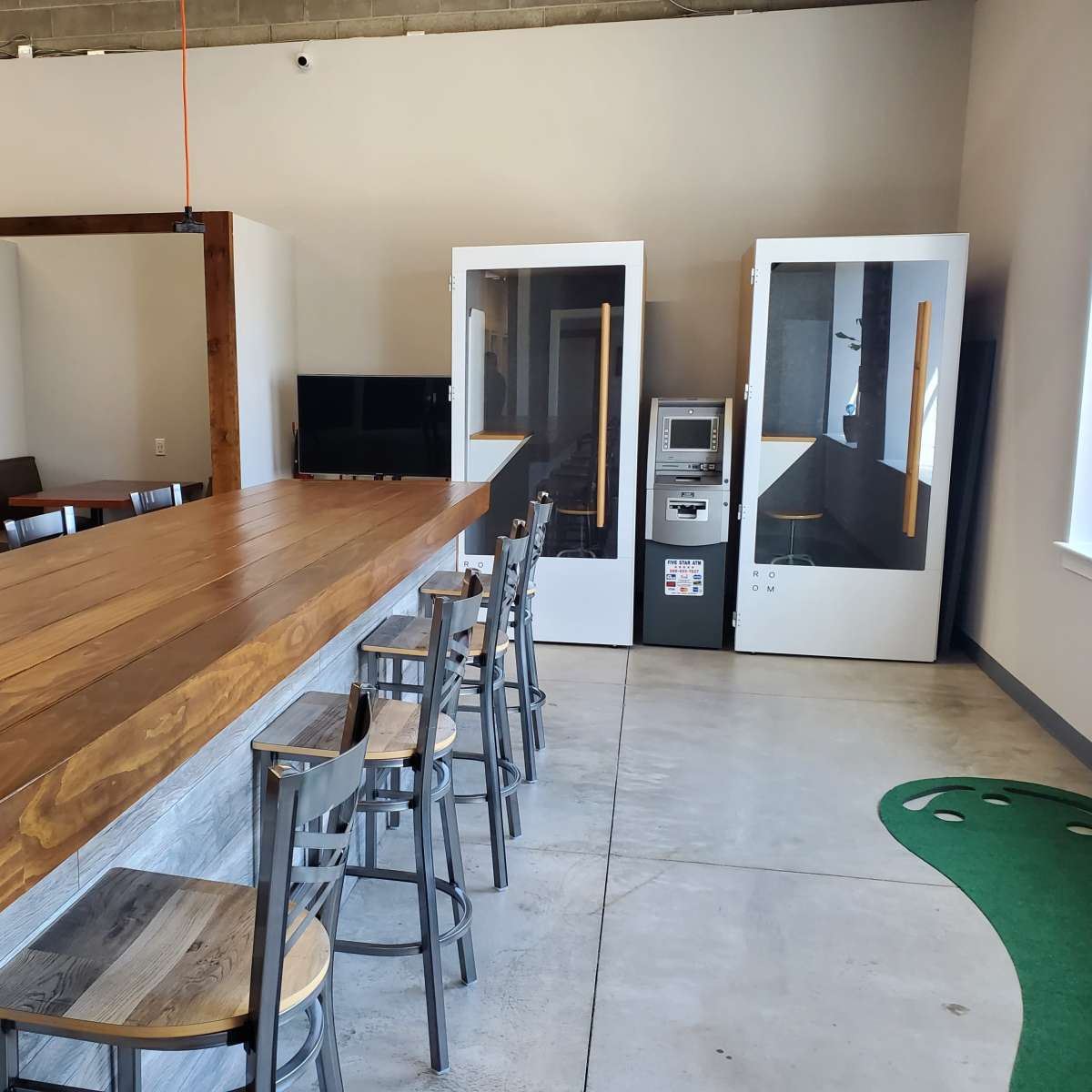 A minimalist workspace with a long wooden countertop, high stools, two soundproof phone booths, a vending machine, and a green putting mat in a well-lit area.