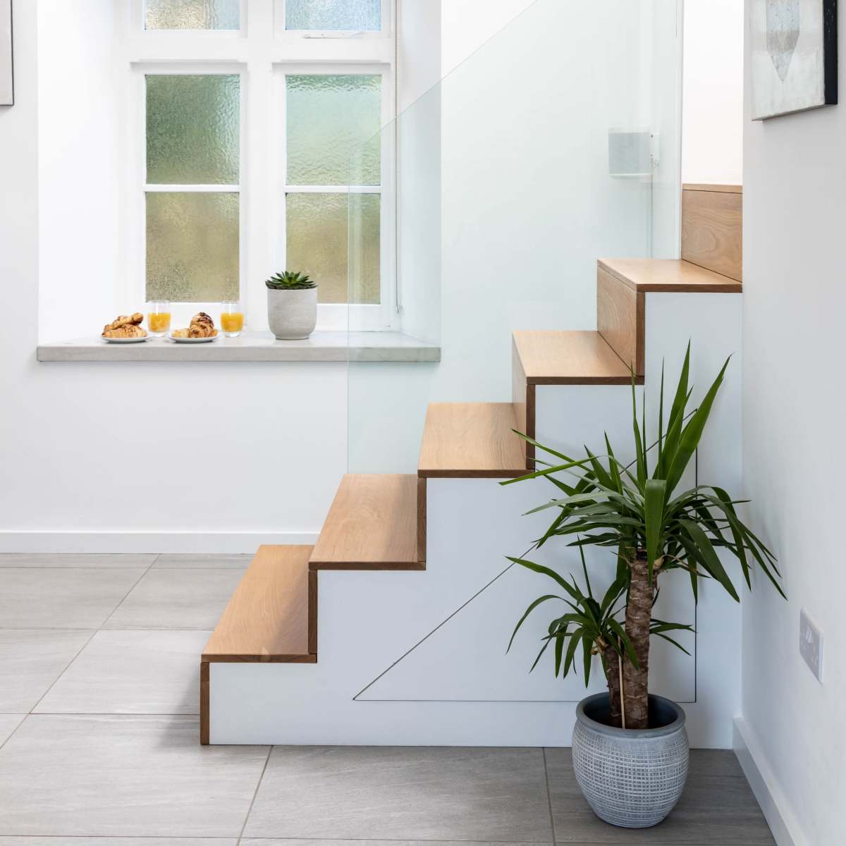 A modern staircase with wooden steps, a glass railing, a potted plant at the bottom, and a window displaying small plates with snacks and drinks.