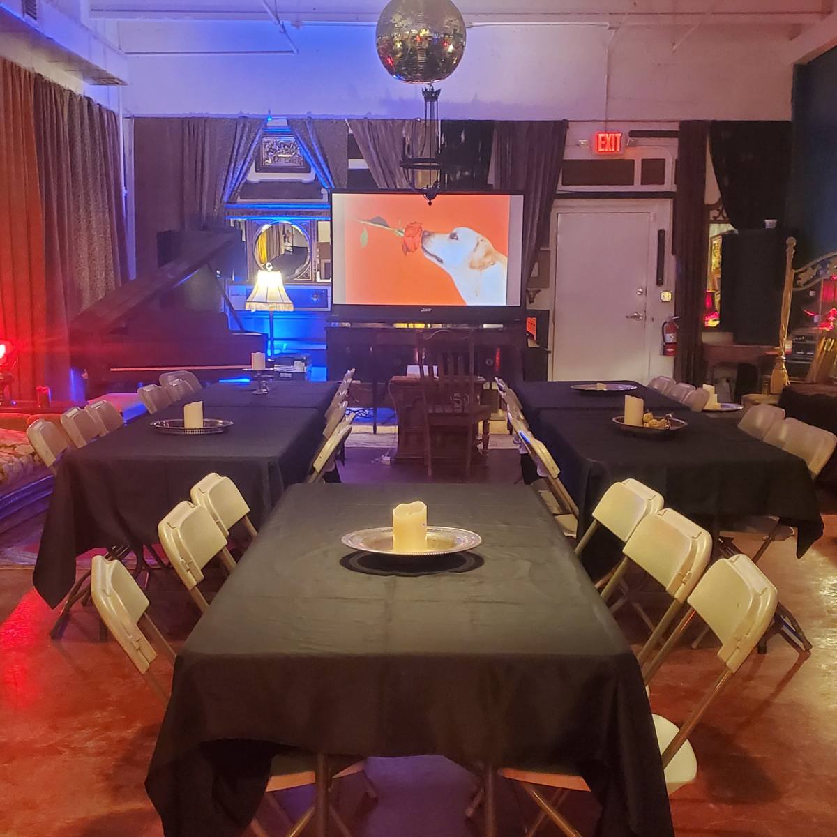 A room with several tables covered in black cloth, arranged in a U-shape, features a screen displaying a film and is illuminated with colorful lights.