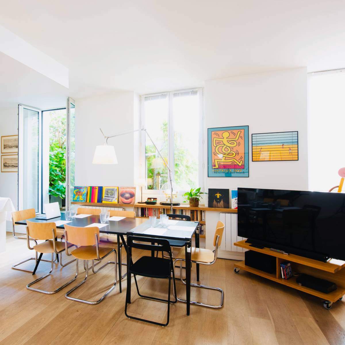 A bright, open living space features a dining area with a table and chairs, a modern TV stand, and large windows leading to a garden.