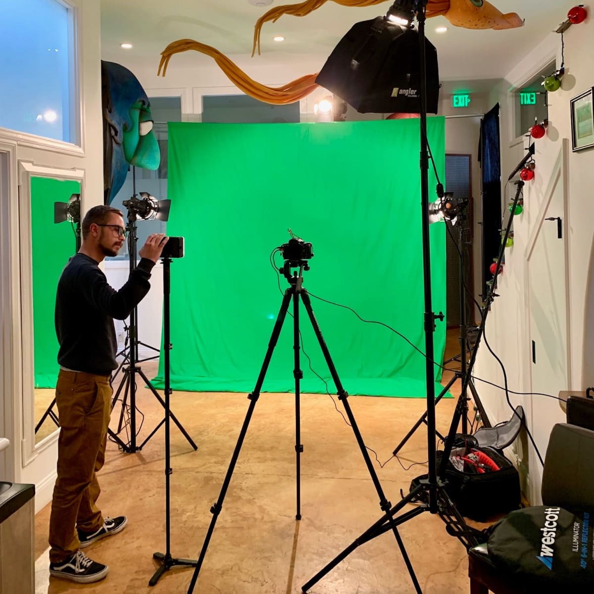 A man sets up a camera in front of a green screen, surrounded by studio lighting and equipment in a recording space.
