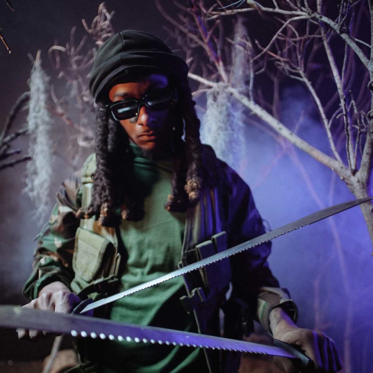 A soldier in camouflage attire holds a saw while standing among bare trees with smoke in the background.