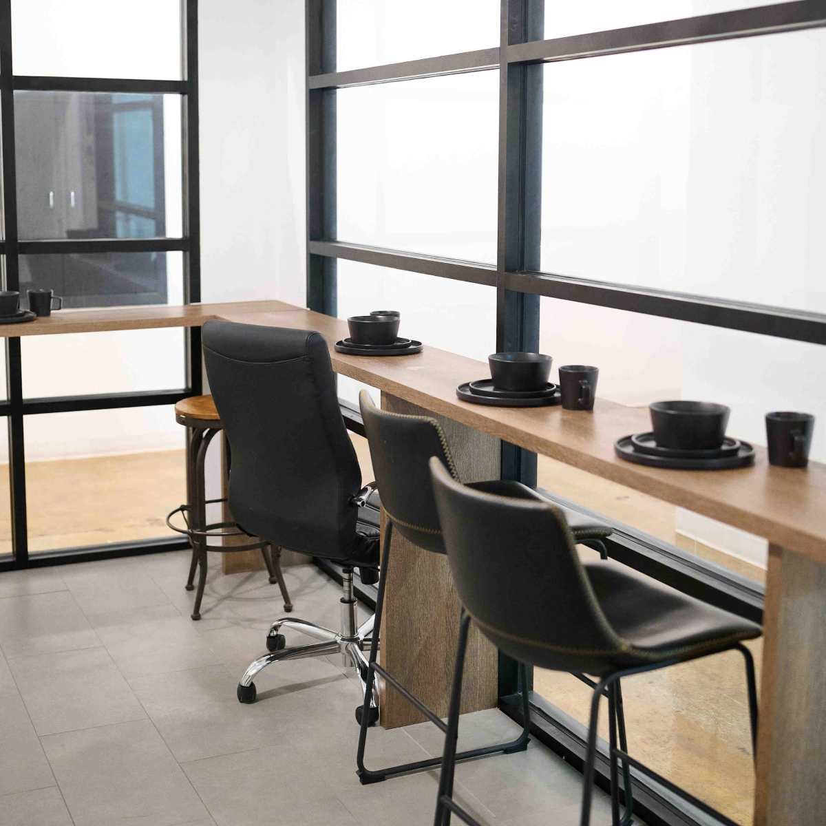 A modern workspace with a long wooden counter, black chairs, and tableware, situated near large glass windows.