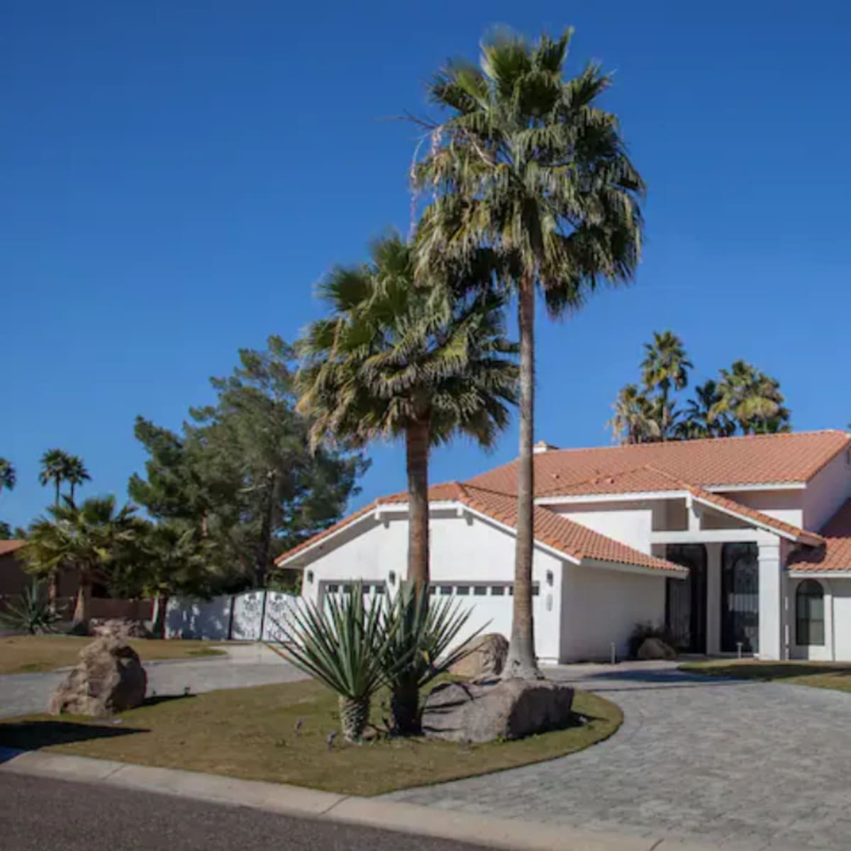 A modern house with a red-tiled roof is situated on a landscaped lot with palm trees and a paved driveway.