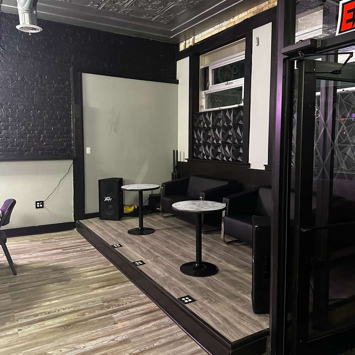 A contemporary seating area with two small round tables and black couches, located in a well-lit space featuring textured walls and a patterned ceiling.