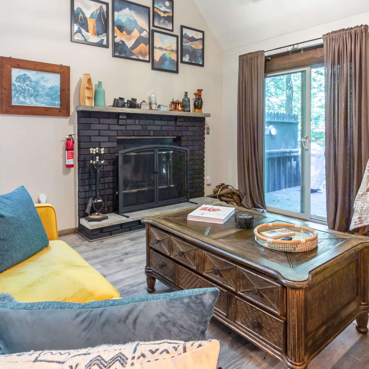 A cozy living room features a yellow sofa, a wooden coffee table, a black fireplace, and large windows opening to a deck.