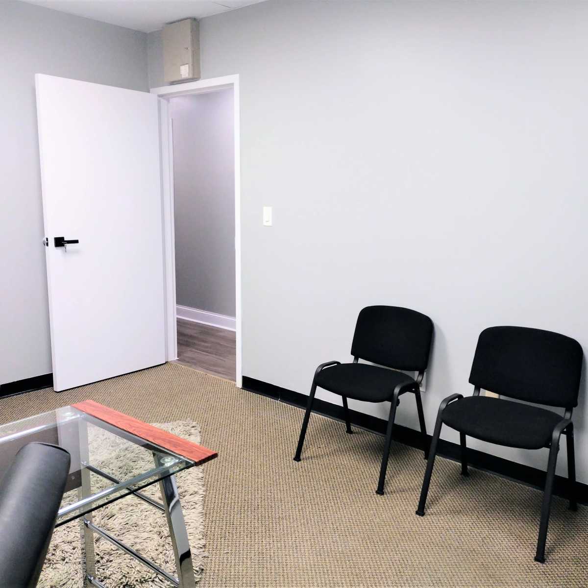 A small, empty room with two black chairs and a glass-topped table, featuring a gray wall and an open door leading to another space.
