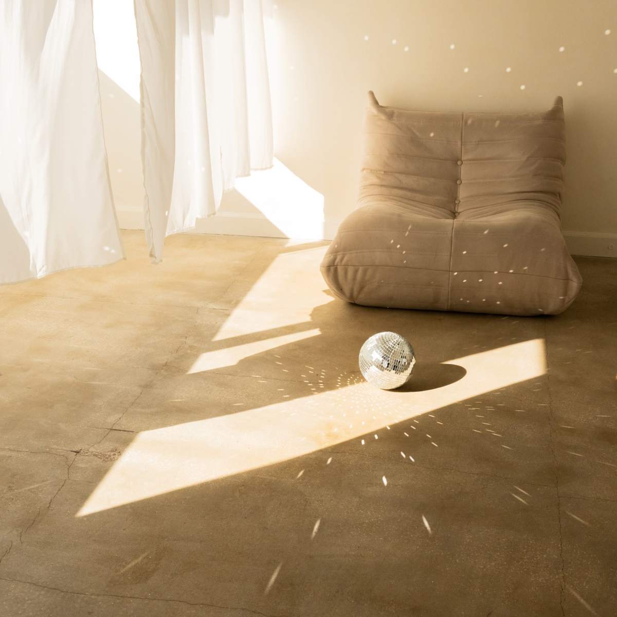 A soft lounge chair sits on a concrete floor, with sunlight filtering through sheer curtains, casting patterns around a reflective disco ball.