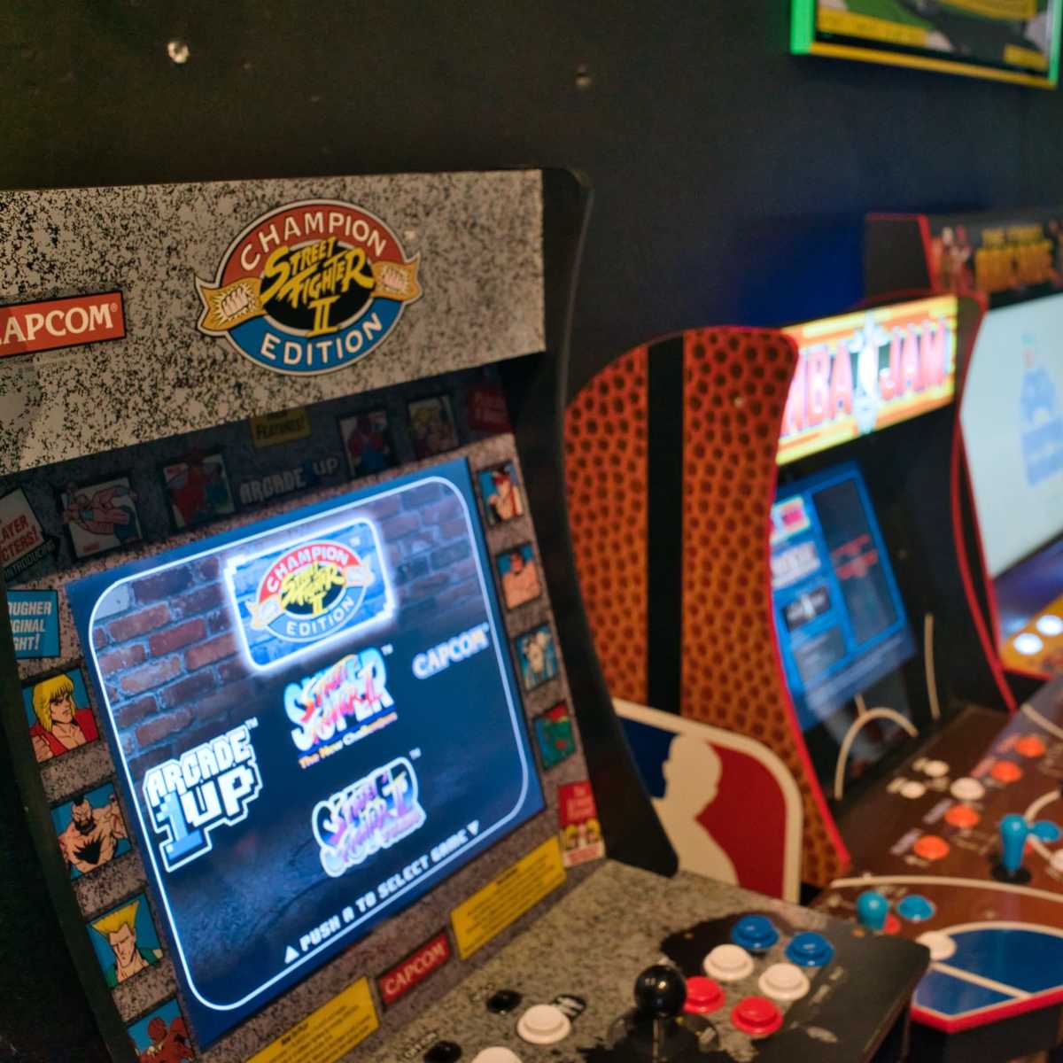 Several vintage arcade game machines, including one labeled "Champion Edition" by Capcom, lined up against a black wall.