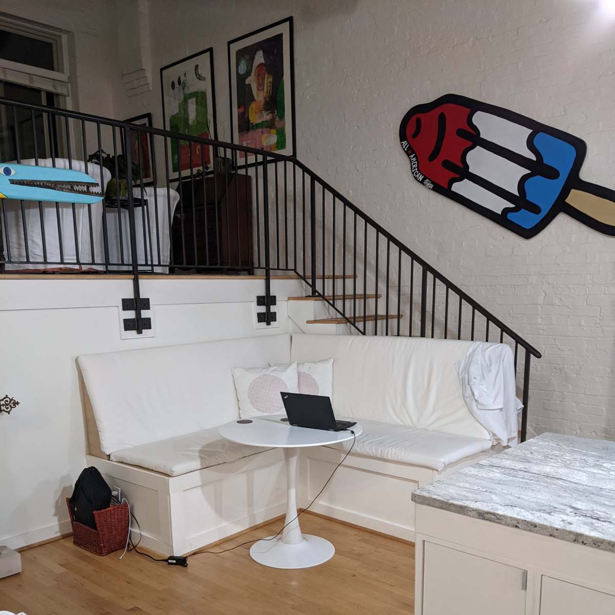 A modern living space featuring a white sectional sofa, a round table with a laptop, a staircase in the background, and colorful wall art.