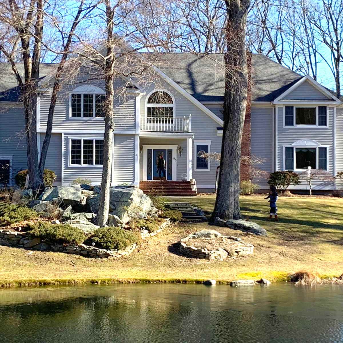 A large house with a gray exterior and multiple windows is situated near a pond, surrounded by trees and landscaped areas.