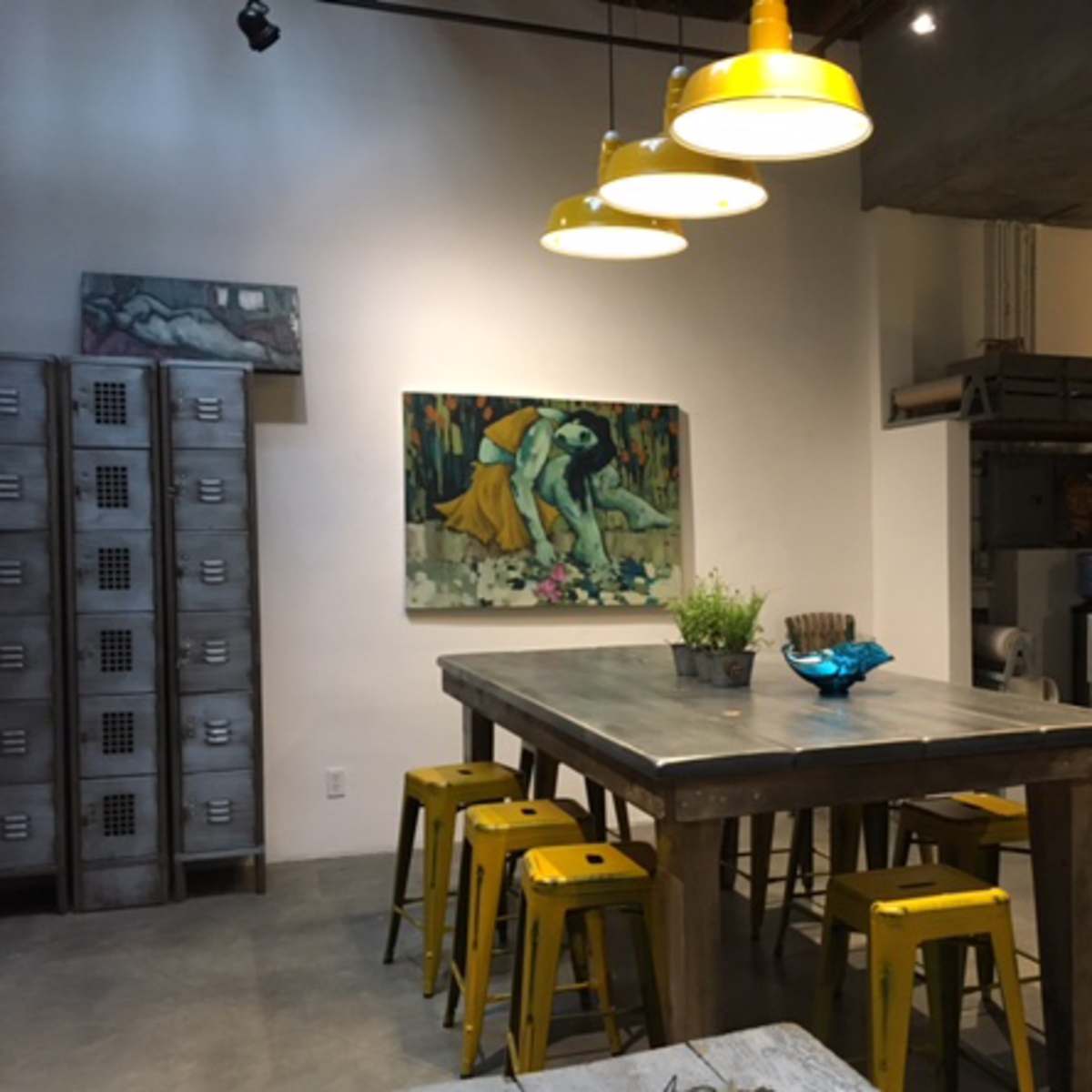 A dining area features a large wooden table surrounded by yellow metal stools, illuminated by pendant lights, with a colorful painting on the wall and shelves holding various kitchen items in the background.