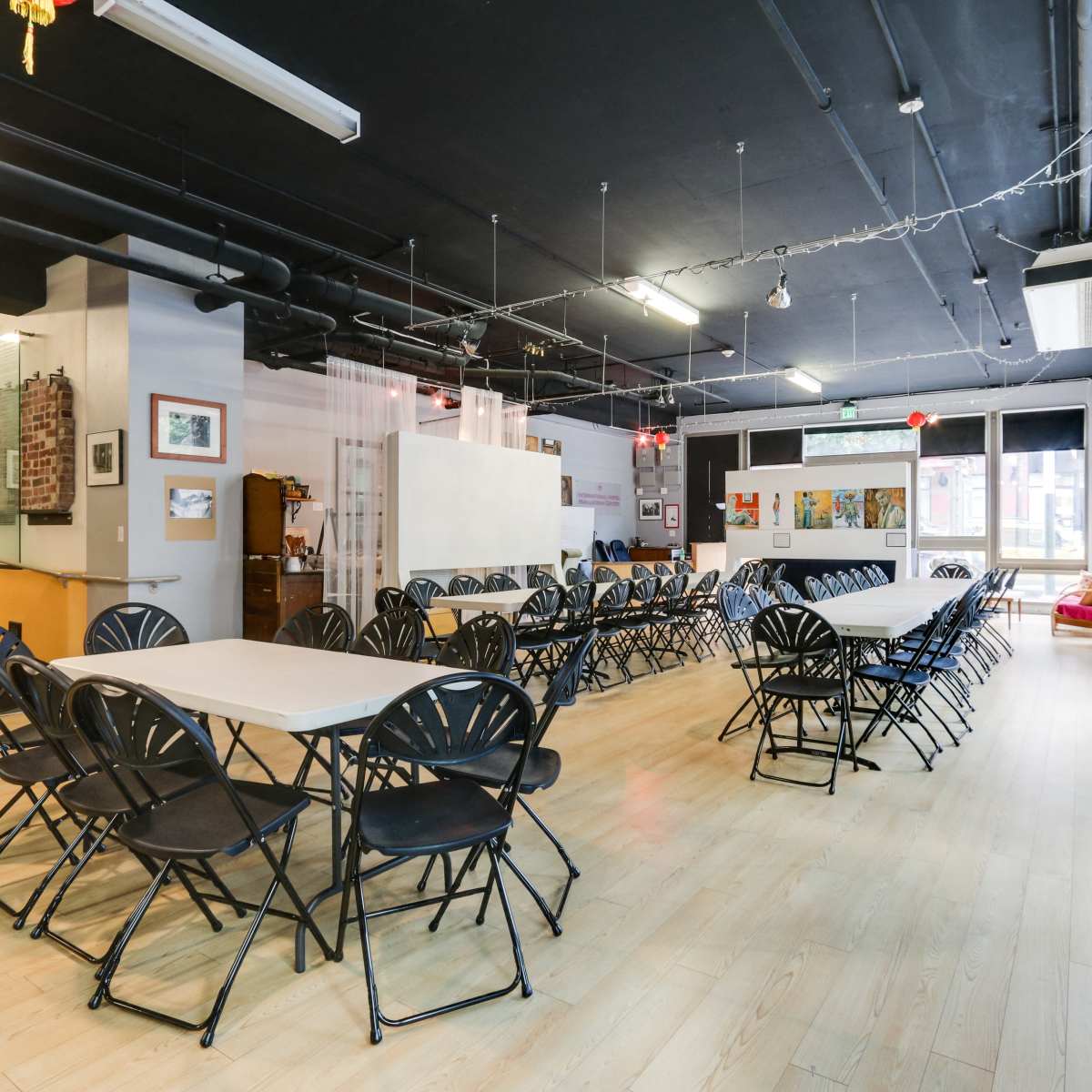 A spacious room with multiple long tables and black folding chairs arranged in rows, illuminated by overhead lights and featuring large windows.