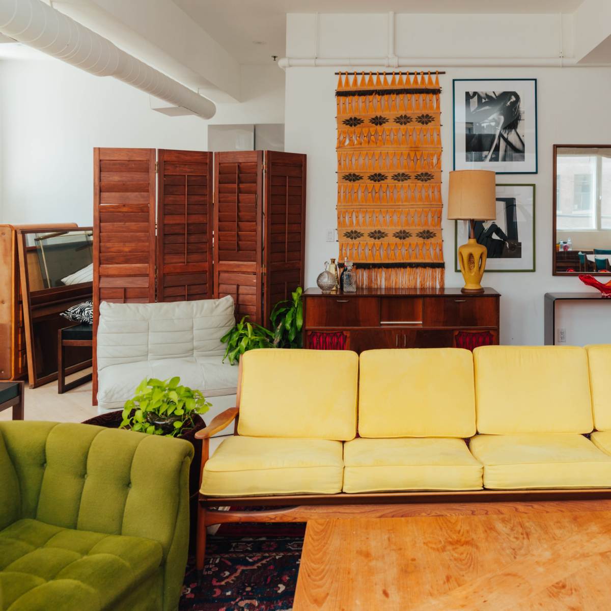 A brightly lit living room features a yellow sofa, a green chair, wooden furniture, and framed artwork on the walls, with plants adding touches of greenery.