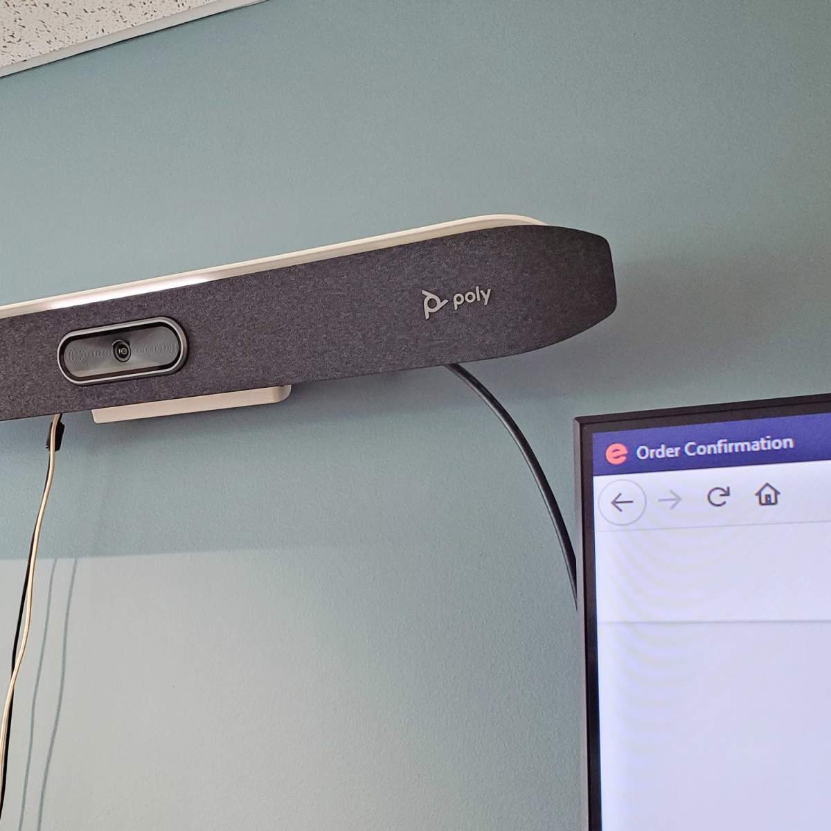A Poly video conferencing device is mounted on a wall above a computer screen displaying an order confirmation.