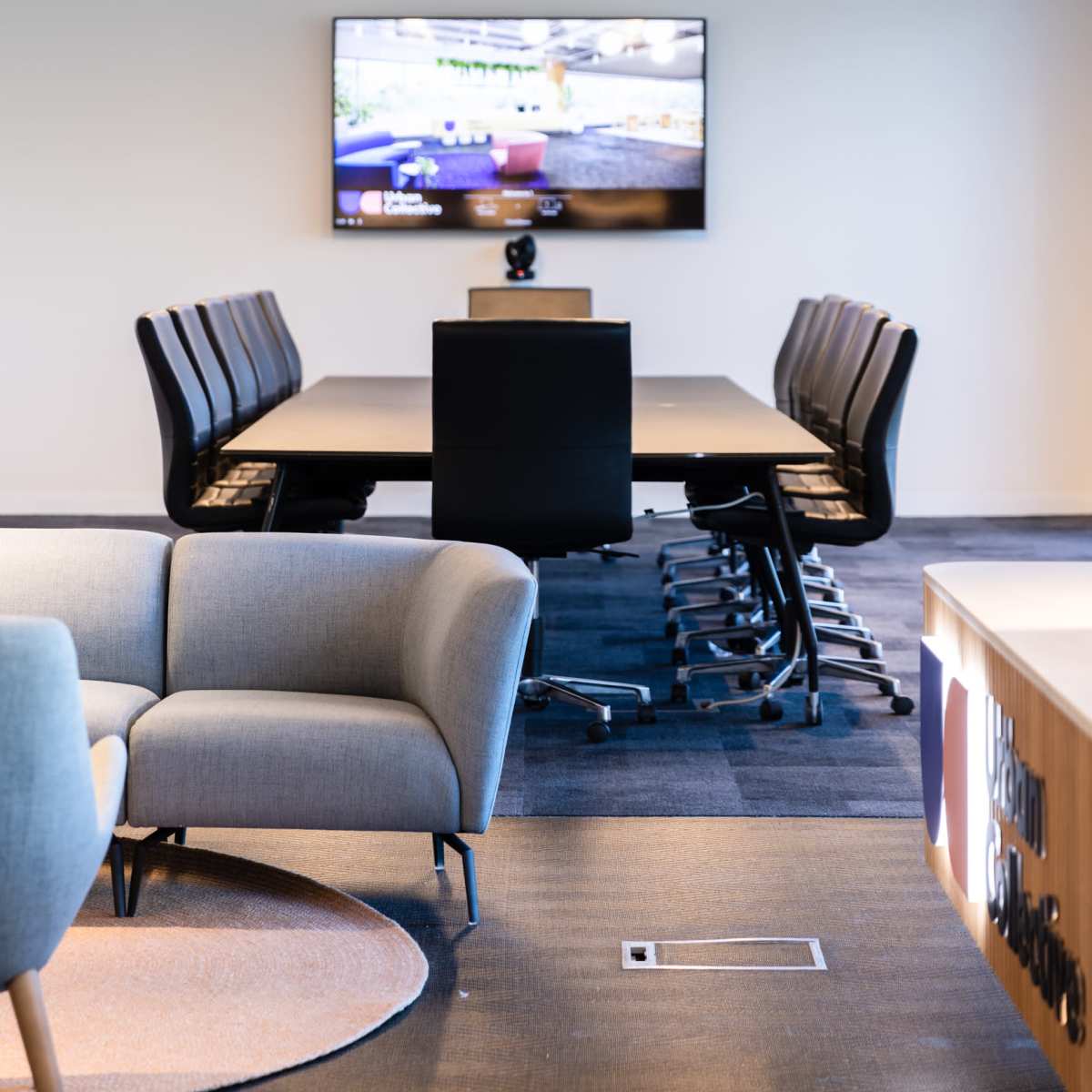 A conference room features a large table surrounded by office chairs, with a wall-mounted television displaying a video game.