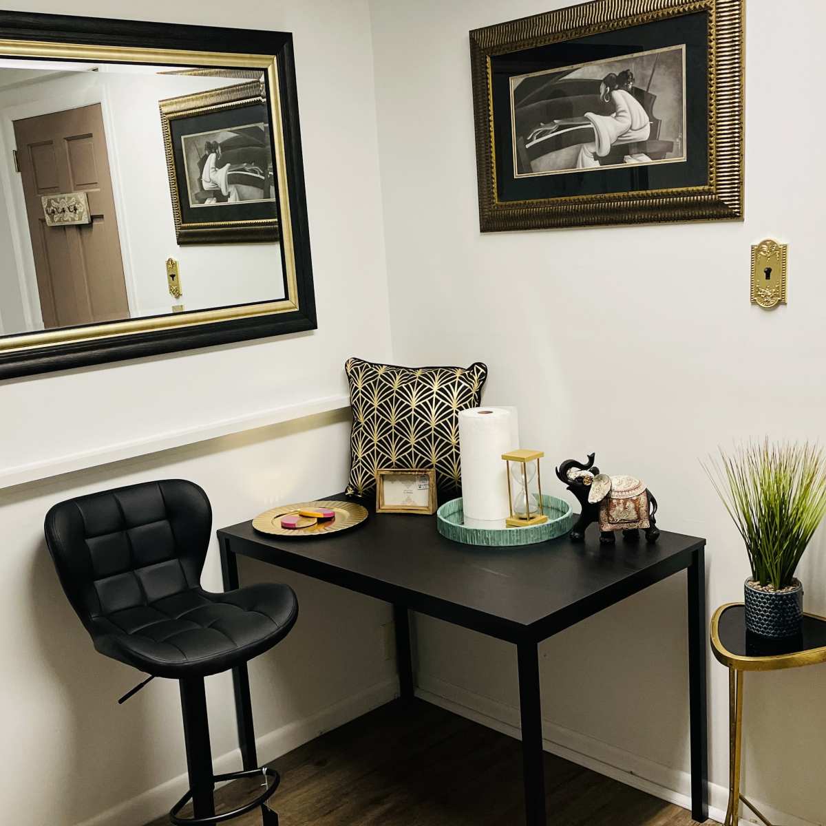 A small corner of a room featuring a black desk with a stool, decorative items on the desk, and framed artwork on the walls.