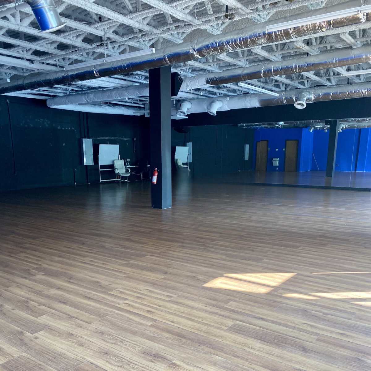 A spacious empty room with wooden flooring and black walls, featuring a few pieces of furniture in the background.